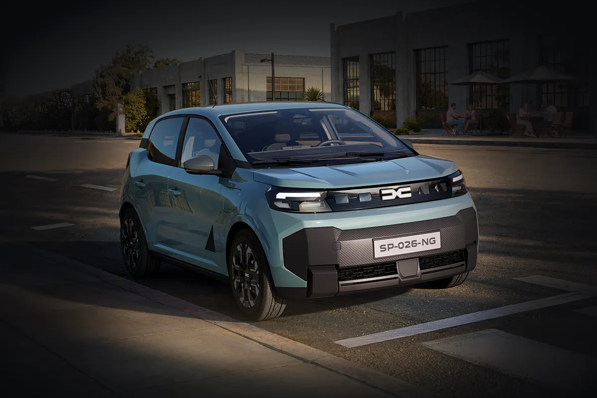 Hellblauer Dacia Spring der neuen Generation von schräg vorne am Straßenrand vor einer Industriehalle mit Café-Terrasse, die Umgebung abgedunkelt, das Auto im warmen Licht.