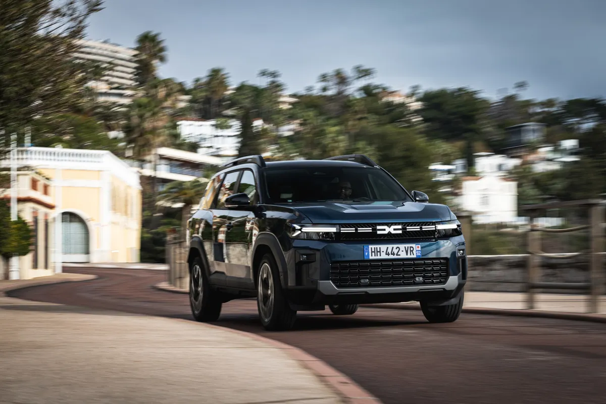 Dunkler Dacia Bigster von schräg vorne in voller Fahrt auf einer sonnigen Straße zwischen hellen Villen und Bäumen.