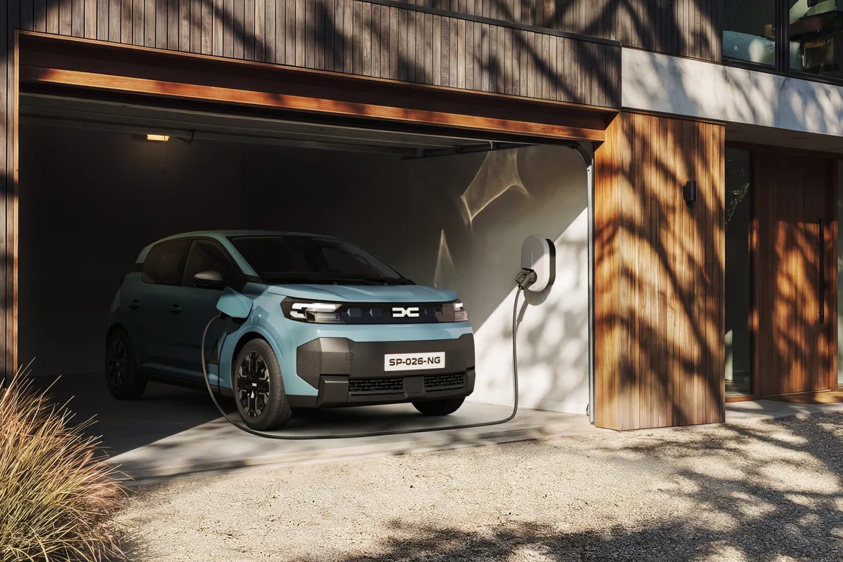 Hellblauer Dacia Spring der neuen Generation von schräg vorne in einer offenen, holzverkleideten Garage, über ein Kabel an einer Wandladestation angeschlossen.