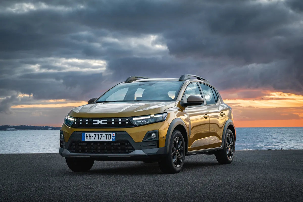 Goldgelber Dacia Sandero Stepway mit schwarzer Dachreling von schräg vorne an einer Uferpromenade bei Sonnenuntergang unter dunklen Wolken.