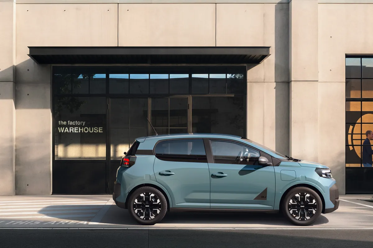 Hellblauer Dacia Spring im Seitenprofil vor einer Industriefassade mit großen Fenstern.