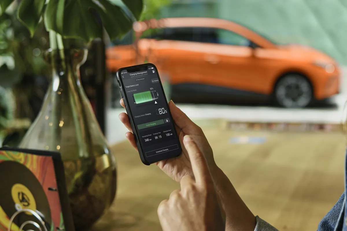 Zwei Hände halten ein Smartphone mit einer Fahrzeug-App, die einen grünen Akku zeigt; dahinter steht unscharf ein oranger MG4.
