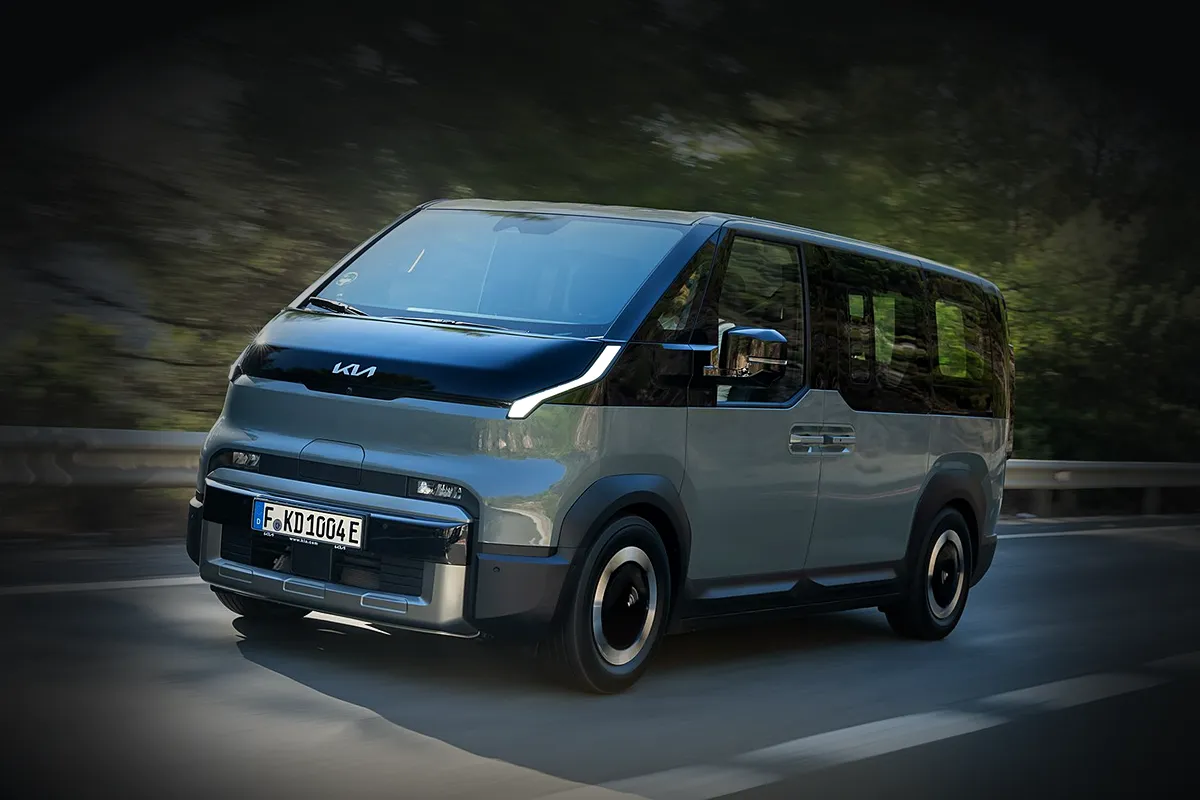 Graugrüner Kia PV5 Passenger von schräg vorne in Fahrt auf einer Landstraße, der Hintergrund mit Bäumen und Leitplanke abgedunkelt.