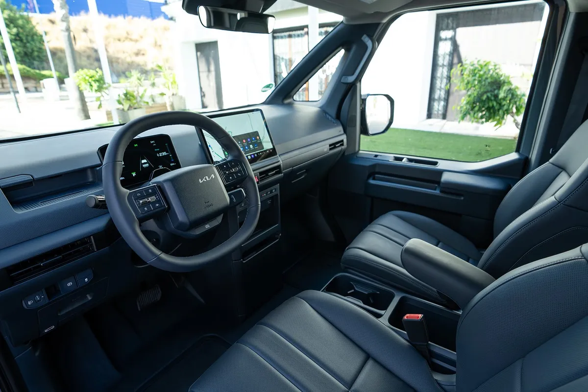 Cockpit eines Kia PV5 von hinten gesehen: Zweispeichenlenkrad, kleines Fahrerdisplay, großer Touchscreen in der Mitte und dunkle Vordersitze.