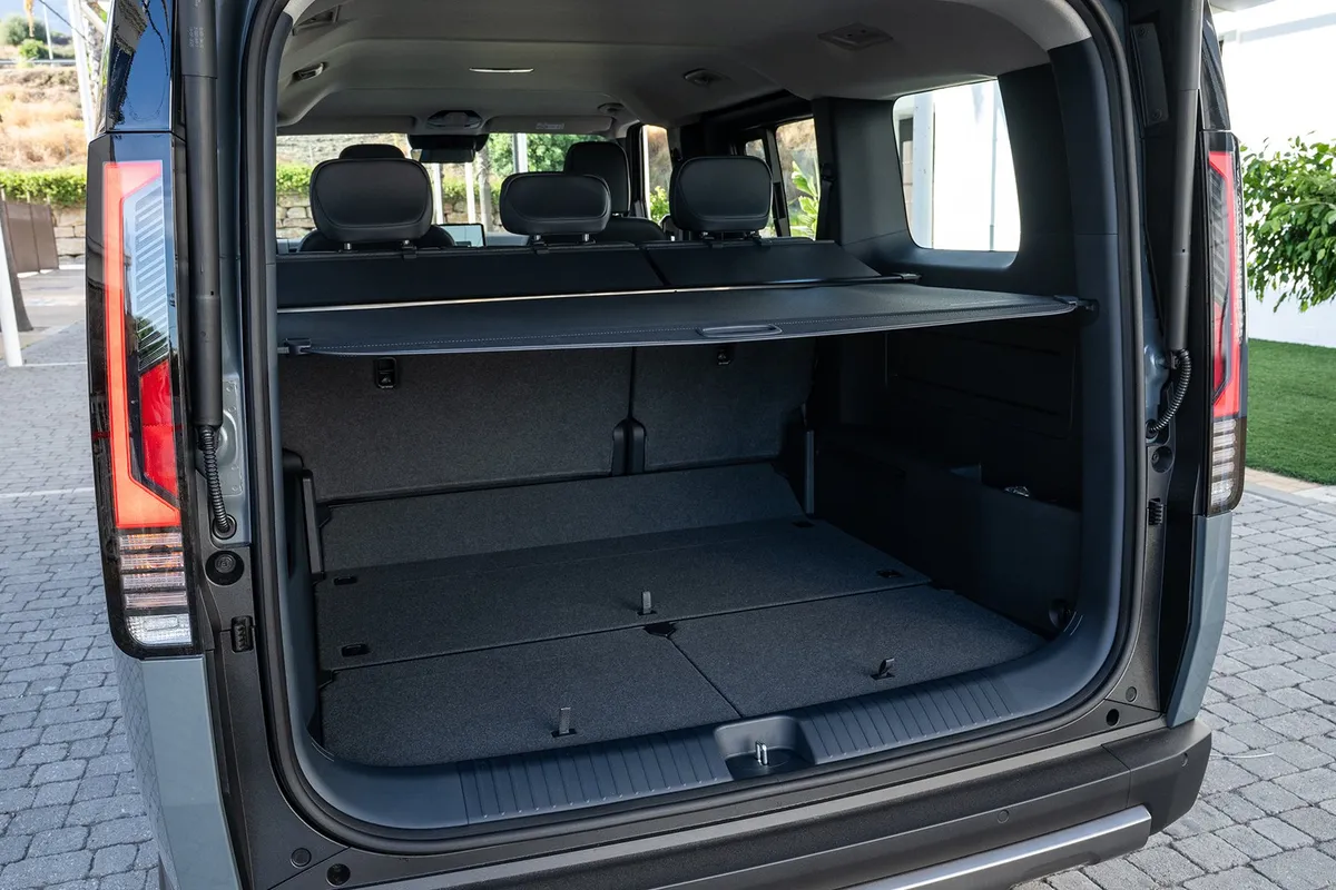 Geöffnete Heckklappe eines Kia PV5 Passenger mit aufrechter Rückbank, Laderaumabdeckung und ebenem Kofferraumboden.