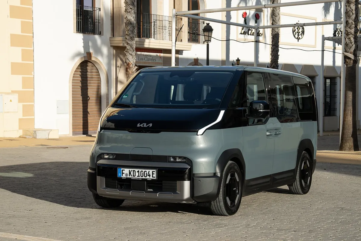 Graugrüner Kia PV5 Passenger von schräg vorne, geparkt vor einem hellen Gebäude mit Balkonen.