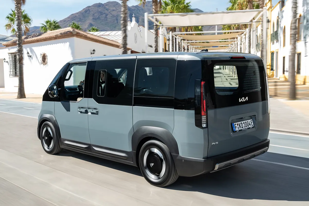 Kia PV5 Passenger von schräg hinten auf einer Straße zwischen weißen Häusern.