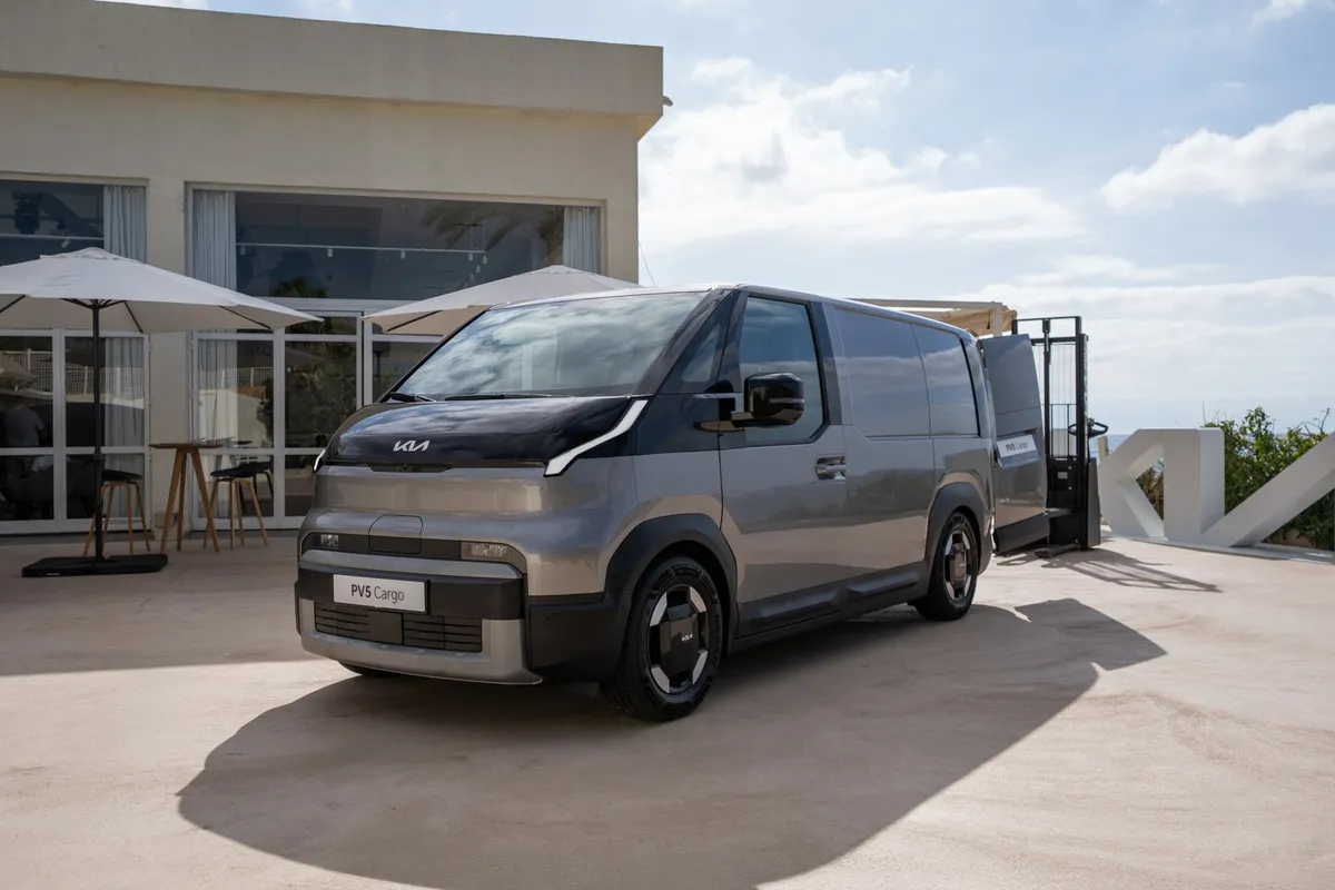 Grauer Kia PV5 Cargo von schräg vorne rechts vor einem modernen Gebäude mit Sonnenschirmen.