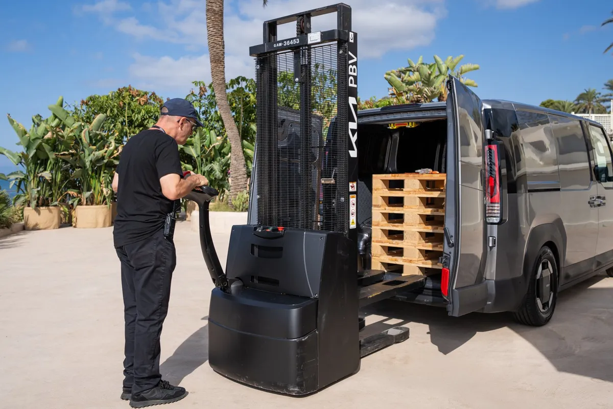 Ein Mann lädt mit einem elektrischen Hubwagen Holzpaletten in den Kia PV5 Cargo mit geöffneten Hecktüren.