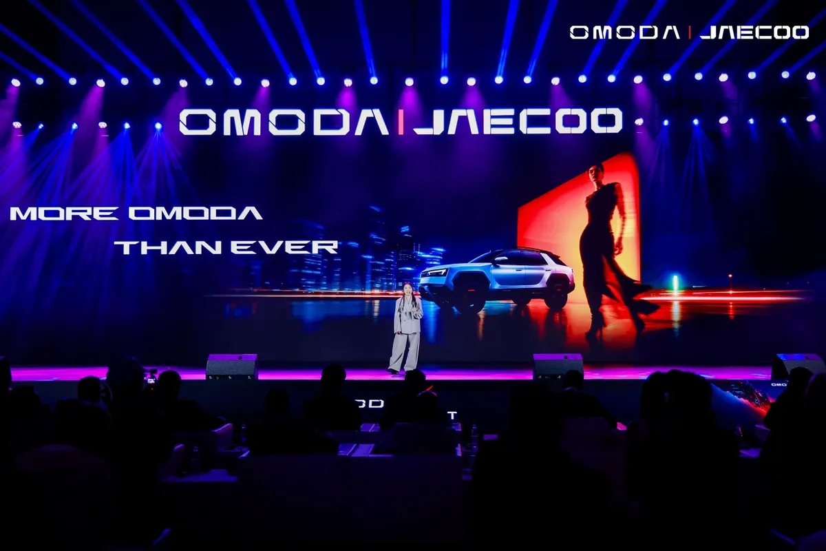 Bühne mit großer Leinwand, auf der der Omoda X zu sehen ist, davor ein Präsentator und Zuschauer.