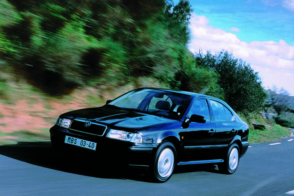 Dunkler Škoda Octavia der ersten Generation fährt auf einer Landstraße mit grünen Bäumen, von schräg vorne fotografiert.