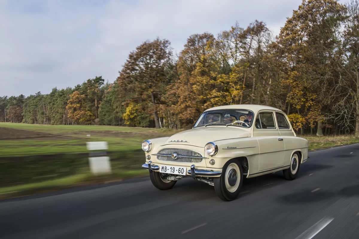 Beiger Škoda Octavia aus den 1960er-Jahren mit runden Scheinwerfern auf einer Landstraße vor herbstlichem Wald.