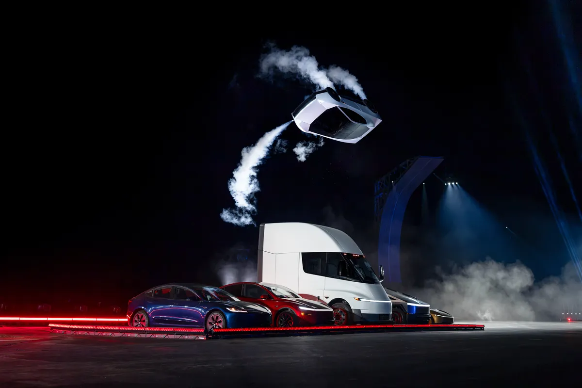 Veranstaltungsszene mit einem schwebenden silbernen Tesla Roadster über einer Bühne mit weißem Sattelschlepper, mehreren Autos, Rauch und roten Lichtstreifen.