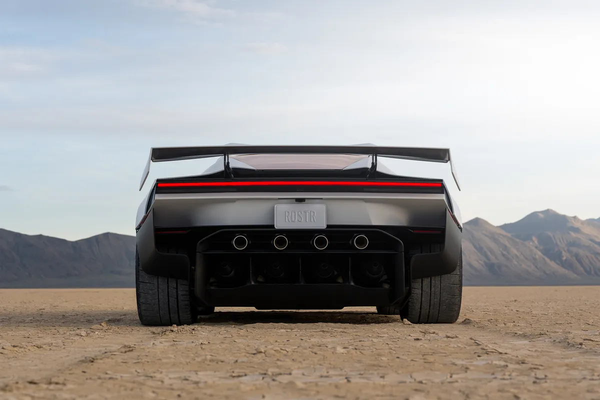 Gerade Heckansicht eines silbernen Tesla Roadster mit großem Heckflügel und vier runden Hecköffnungen auf trockenem Wüstenboden vor einer Bergkette.