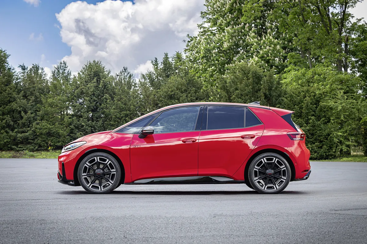 Roter VW ID.3 GTI im Seitenprofil auf einer Asphaltfläche vor Bäumen, mit schwarzen Außenspiegeln und zweifarbigen geometrischen Felgen.