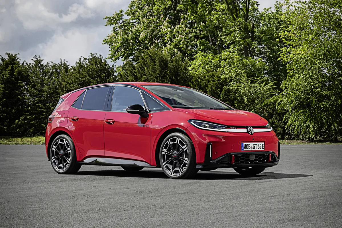 Roter VW ID.3 GTI von schräg vorne auf einer Asphaltfläche vor dichtem Grün, mit großem unteren Lufteinlass und geometrischen Felgen.