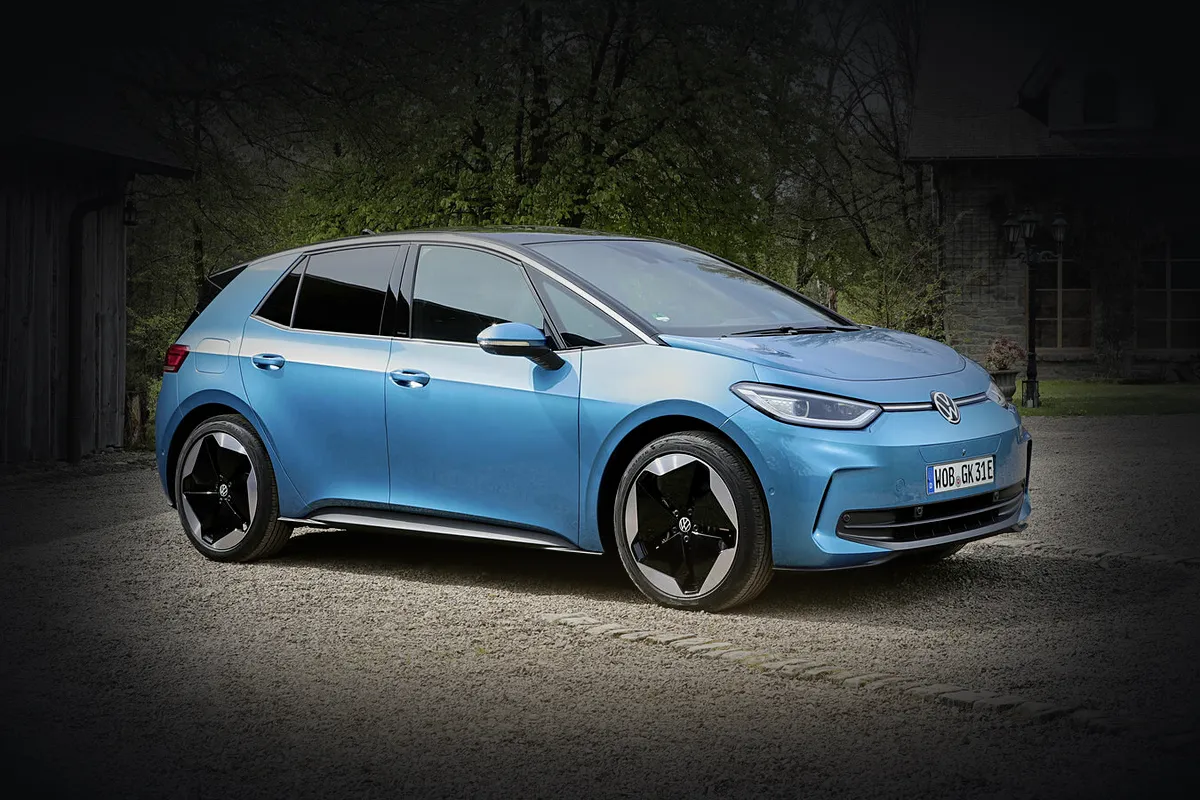 Hellblauer VW ID.3 der zweiten Generation von schräg vorne auf einem Kiesplatz, die Umgebung abgedunkelt, das Auto im hellen Licht.