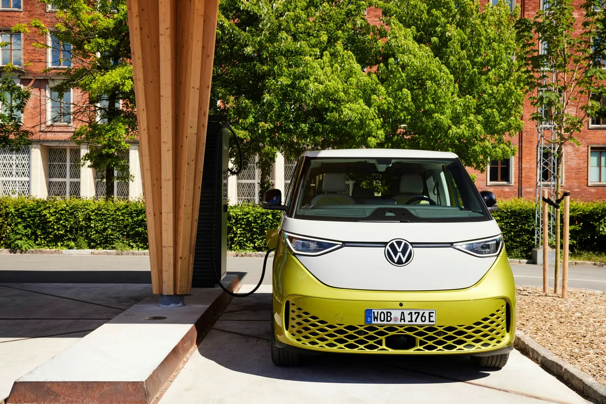 Gelb-weißer VW ID. Buzz frontal an einer Ladestation unter einem Holzdach vor Backsteinhäusern.