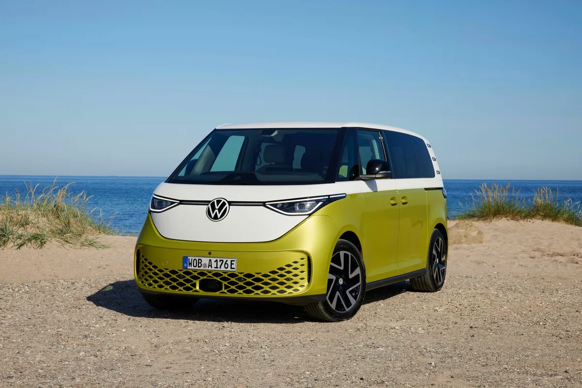 Gelb-weißer VW ID. Buzz von schräg vorne auf sandigem Boden vor Dünen und dem Meer.
